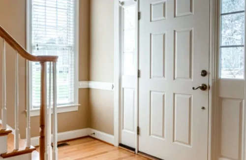 Eine weiße Haustür mit altmodischer Türklinke und kleinen Seitlichen Glasscheiben vor einem Treppenaufgang.