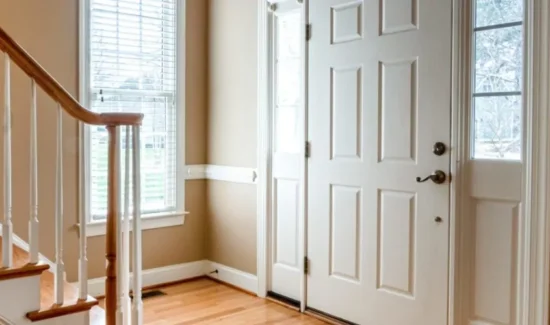 Eine weiße Haustür mit altmodischer Türklinke und kleinen Seitlichen Glasscheiben vor einem Treppenaufgang.