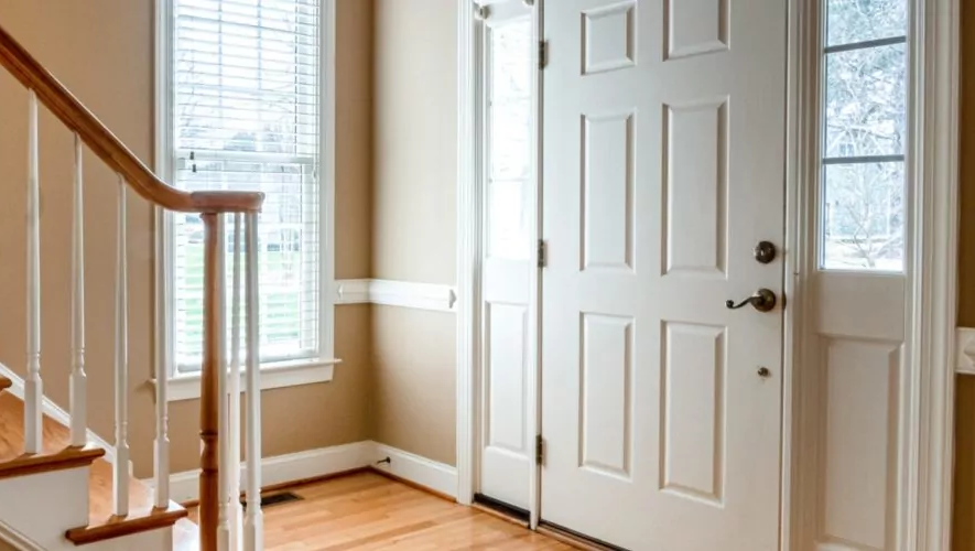 Eine weiße Haustür mit altmodischer Türklinke und kleinen Seitlichen Glasscheiben vor einem Treppenaufgang.