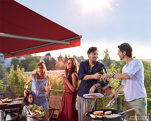 Eine Freundesgruppe beim Grillen auf der Terrasse unter einer roten Markise.