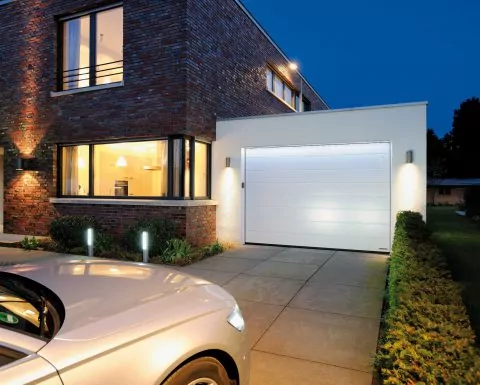 Eine lange Auffahrt aus großen Betonplatten führt zu einem Garagentor in weißer Farbe mit seitlich angebrachten Lampen.