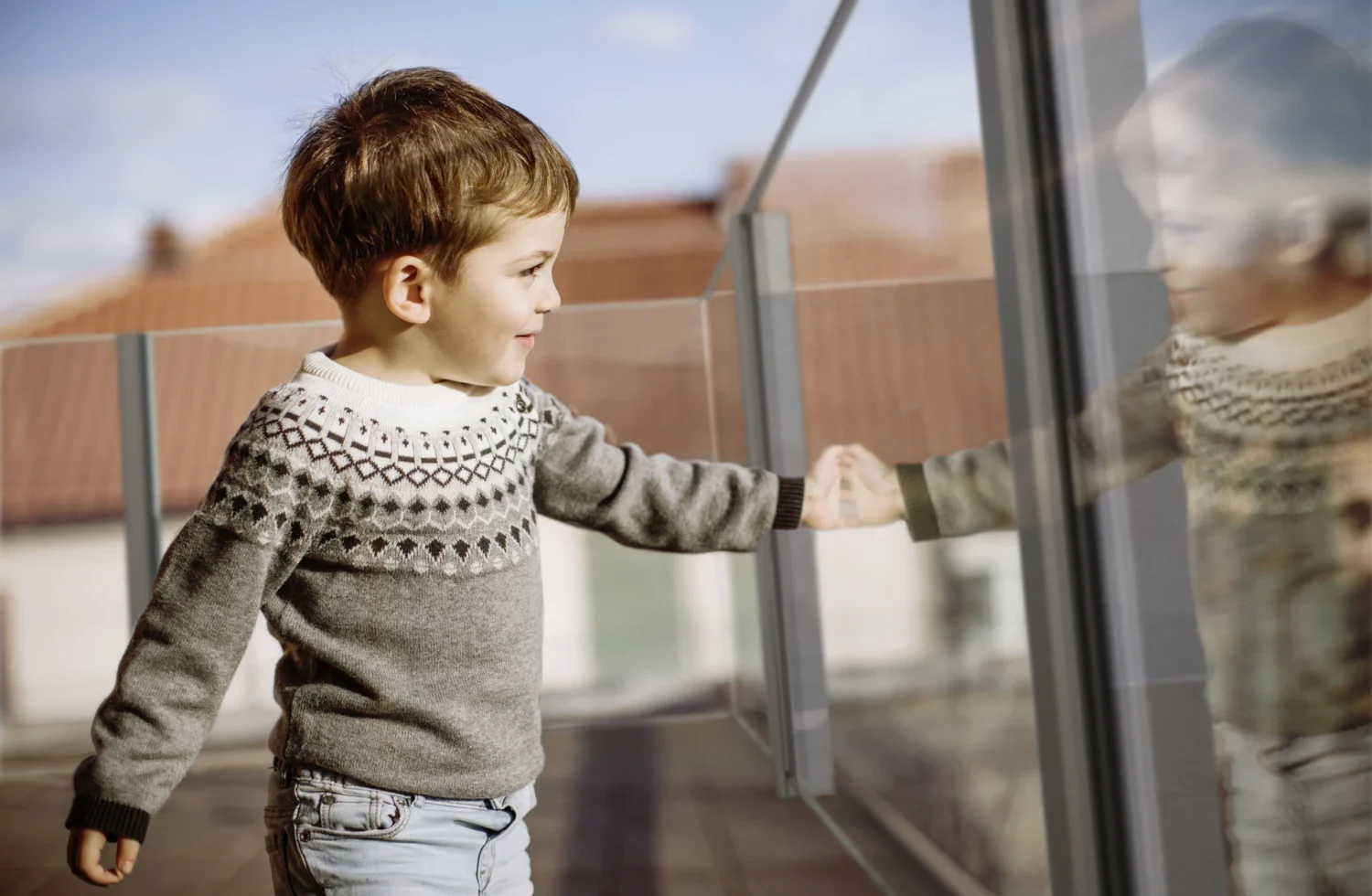 Kind in grauem Pullover betrachtet seine Spiegelung in einer Glaswand